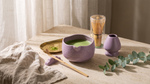 Purple ceramic matcha whisk set on a travertine counter with linen and brass tray