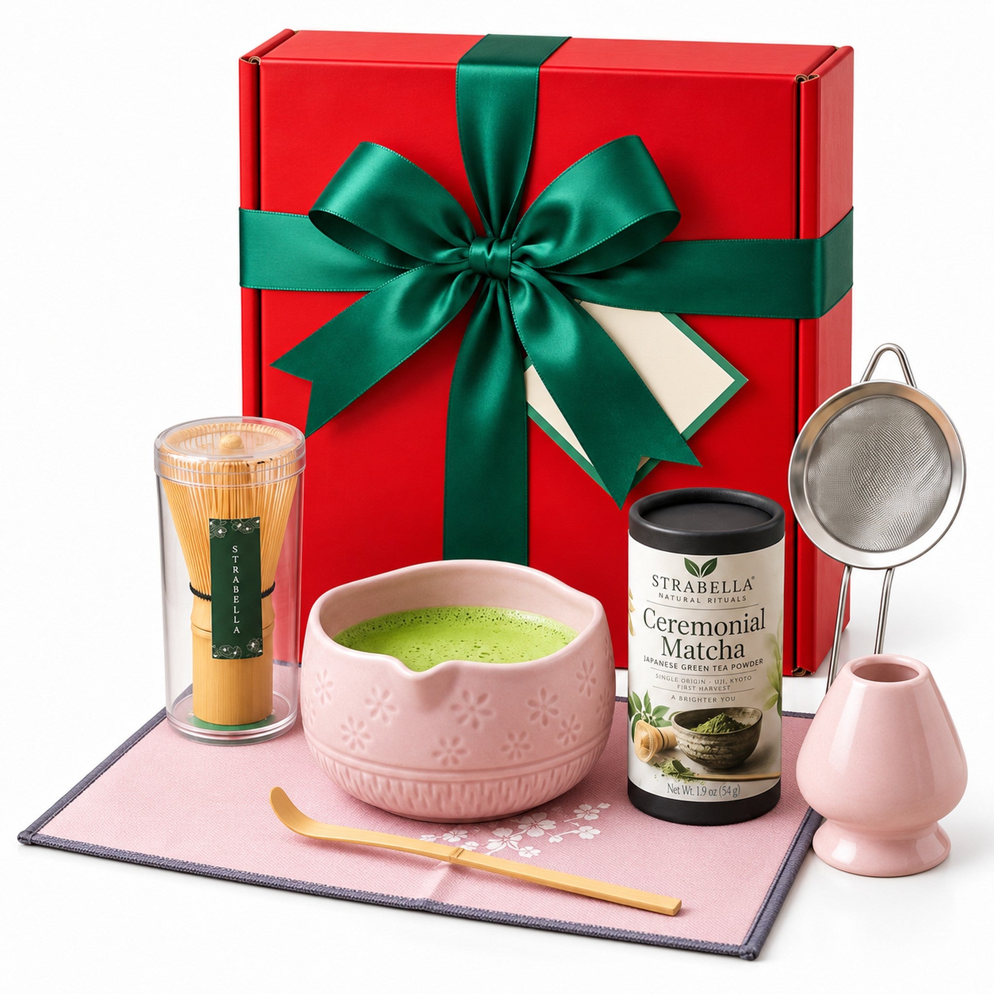 Christmas Matcha Gift Box, Pink: red gift box with a green satin bow standing behind the pink 7-piece matcha whisk set and Ceremonial Matcha tin