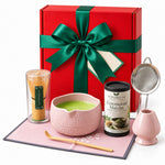 Christmas Matcha Gift Box, Pink: red gift box with a green satin bow standing behind the pink 7-piece matcha whisk set and Ceremonial Matcha tin
