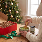 Hands whisking matcha in a ceramic bowl beside the opened red gift box and a Christmas tree