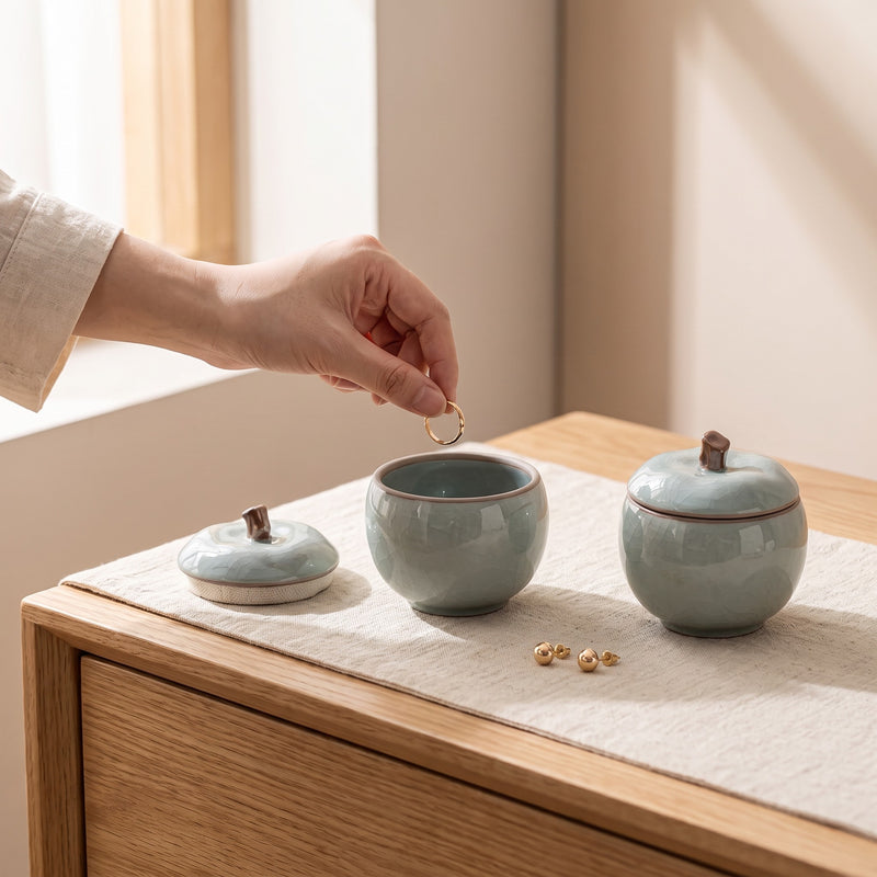 Trinket jar — hand dropping a ring into an open azure apple jar on a dresser